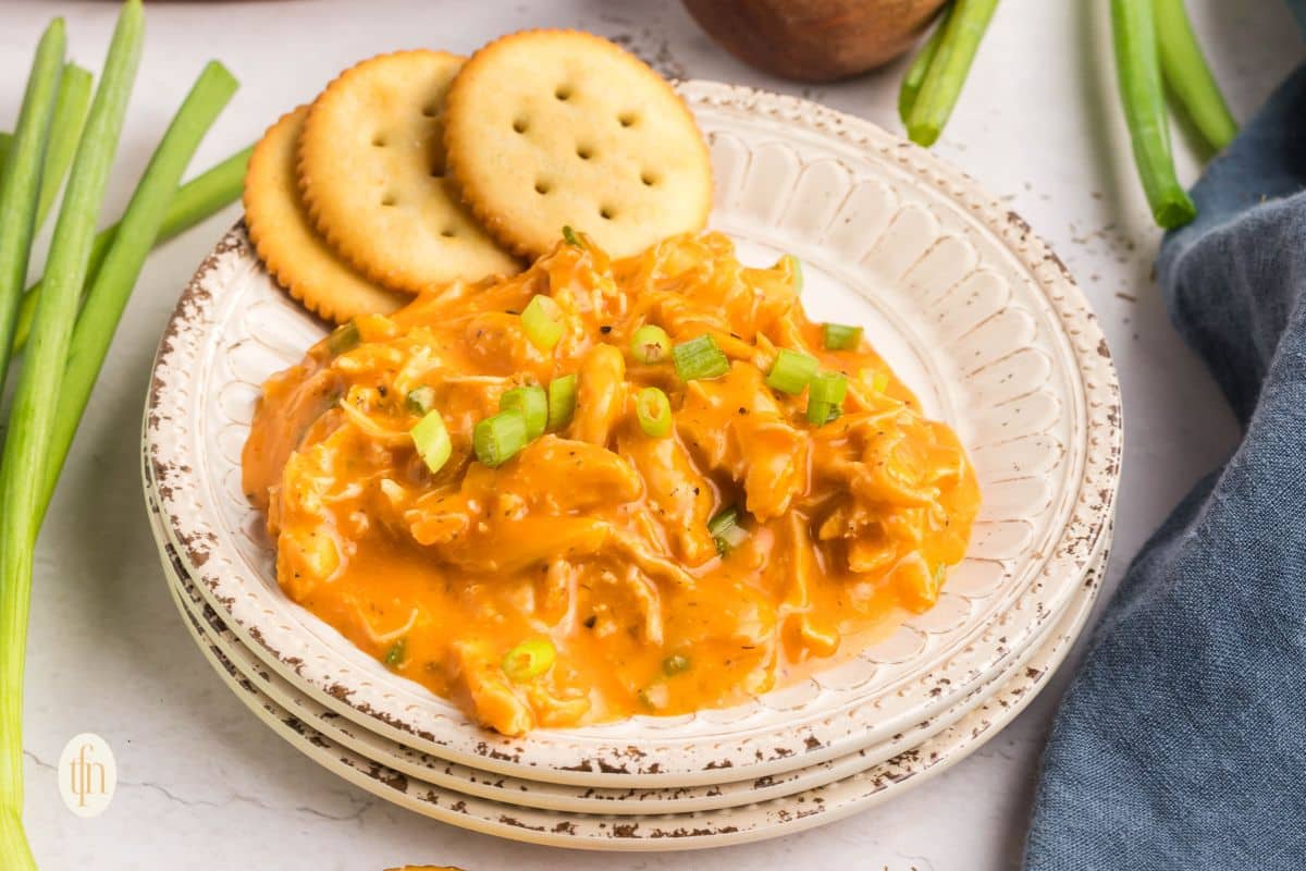 Creamy buffalo chicken dip topped with chopped green onions, served with round crackers on a white plate.