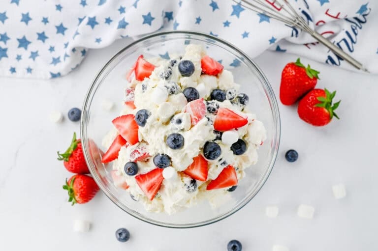 A glass bowl of creamy fruit salad with strawberries, blueberries, and mini marshmallows on a white surface.