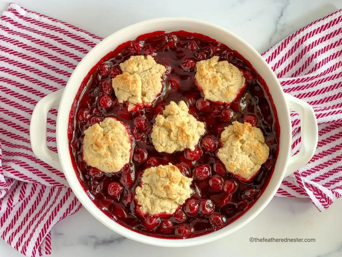 Baked cherry cobbler with six biscuit toppings in a round white dish on a red striped towel.