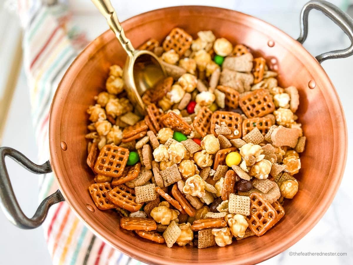 A copper bowl filled with snack mix, including pretzels, popcorn, cereal, and candy, with a scoop inside.