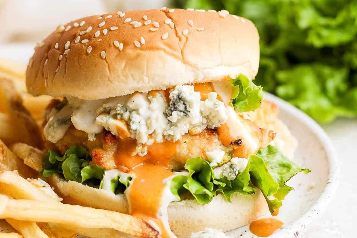Chicken sandwich with lettuce, blue cheese, and sauce, served with French fries on a white plate.