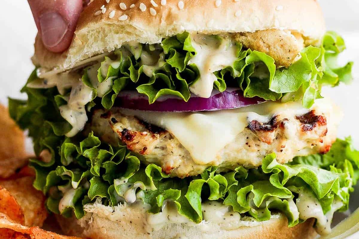 Close-up of a chicken sandwich with lettuce, onion, cheese, and sauce on a sesame bun, held by a hand.