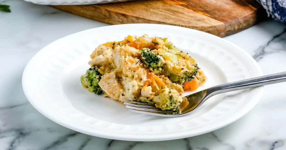 A serving of chicken and broccoli casserole on a white plate with a fork, on a marble countertop.