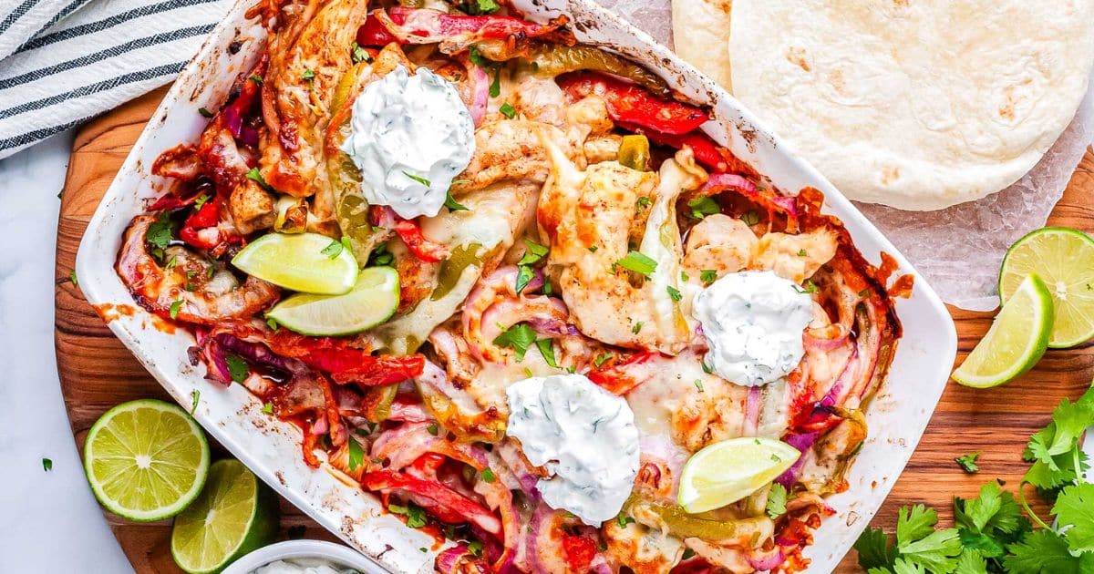 Baked chicken fajitas with peppers, onions, melted cheese, lime wedges, and dollops of sour cream in a dish.