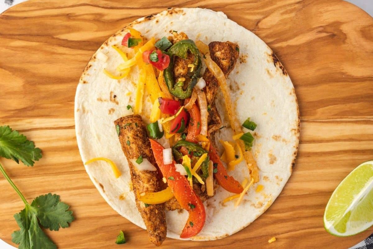 A tortilla with grilled chicken strips, sautéed peppers, onions, and cheese on a wooden board with lime and cilantro.