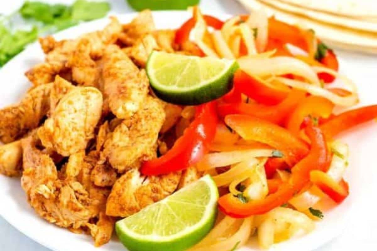 Plate of seasoned chicken strips, sautéed bell peppers and onions, with lime wedges on the side.