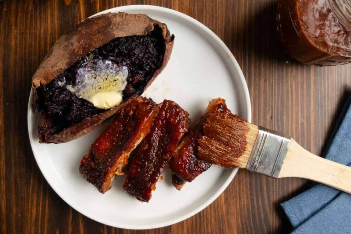 Barbecue ribs with sauce, a baked purple sweet potato with butter, and a basting brush on a white plate.