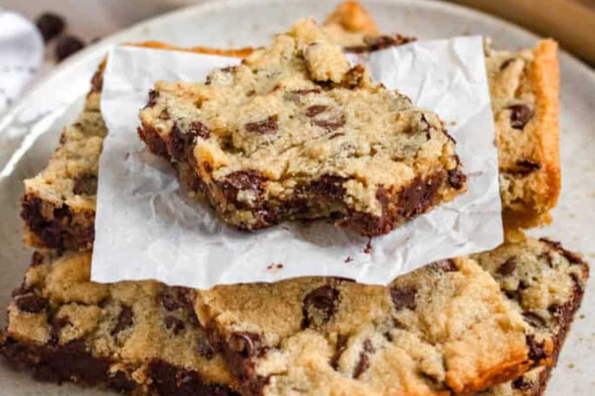 A stack of chocolate chip cookie bars on a plate, with one bar on top partly eaten and resting on parchment paper.