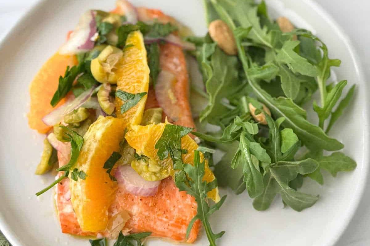 Salmon topped with orange slices, onions, olives, and herbs, served with arugula and almonds on a white plate.
