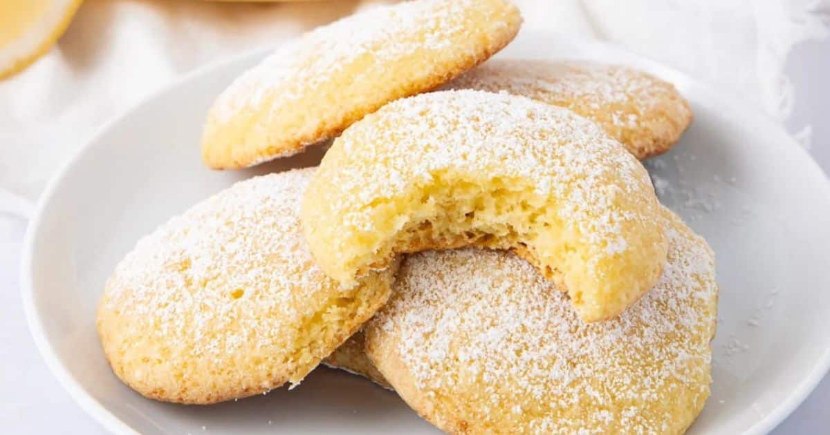 Four powdered sugar cookies on a plate, with one cookie showing a bite taken out of it.