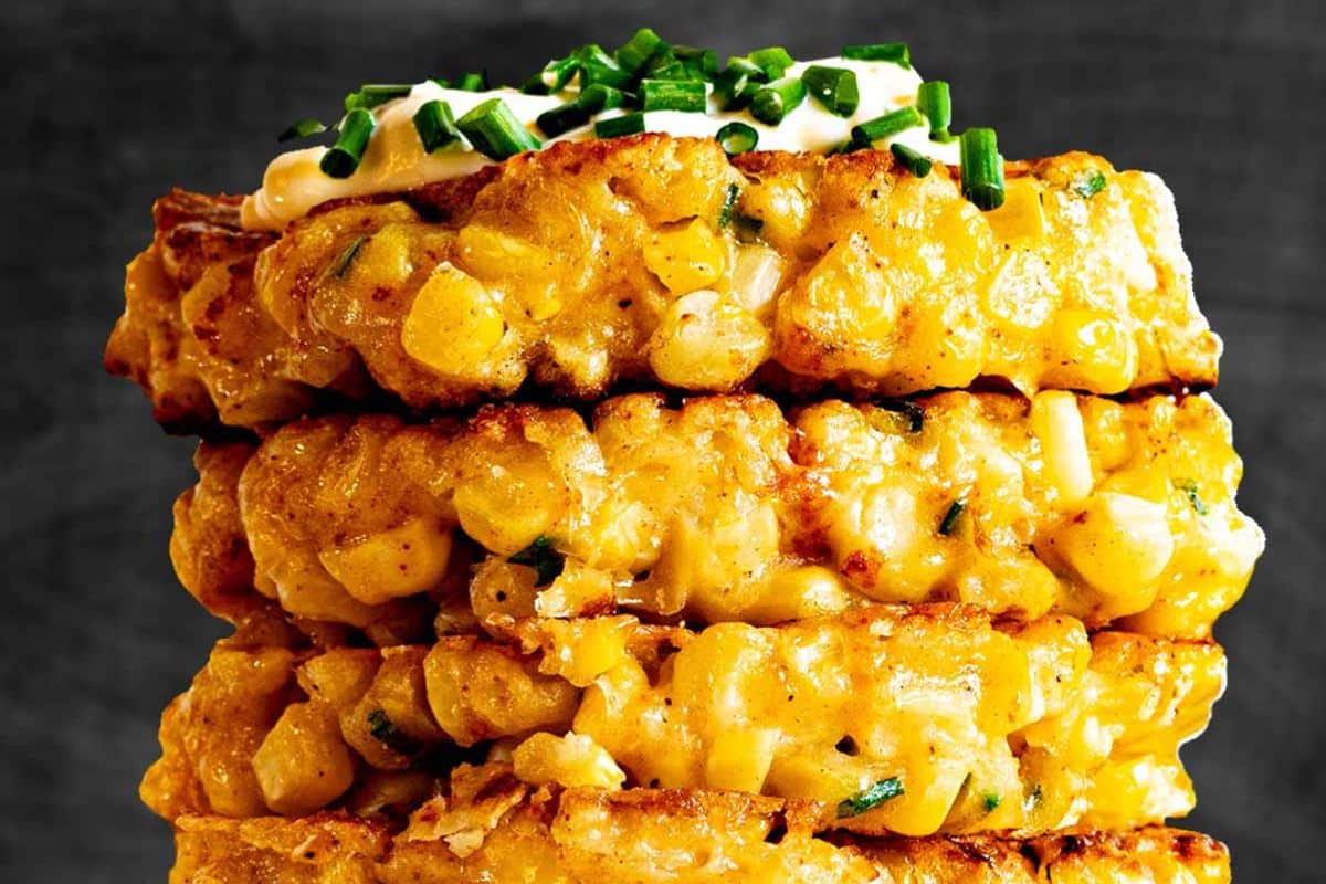 A stack of crispy corn fritters topped with sour cream and chopped chives against a dark background.