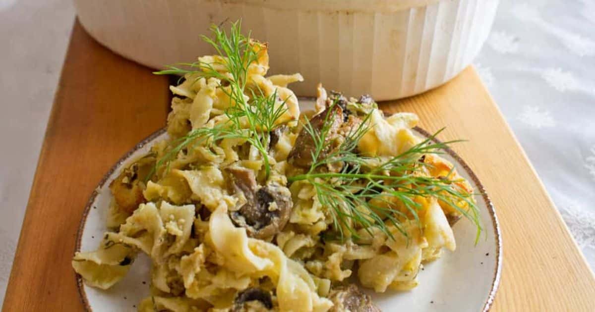 A plate of noodle casserole with mushrooms and herbs, garnished with fresh dill, sits on a wooden board.