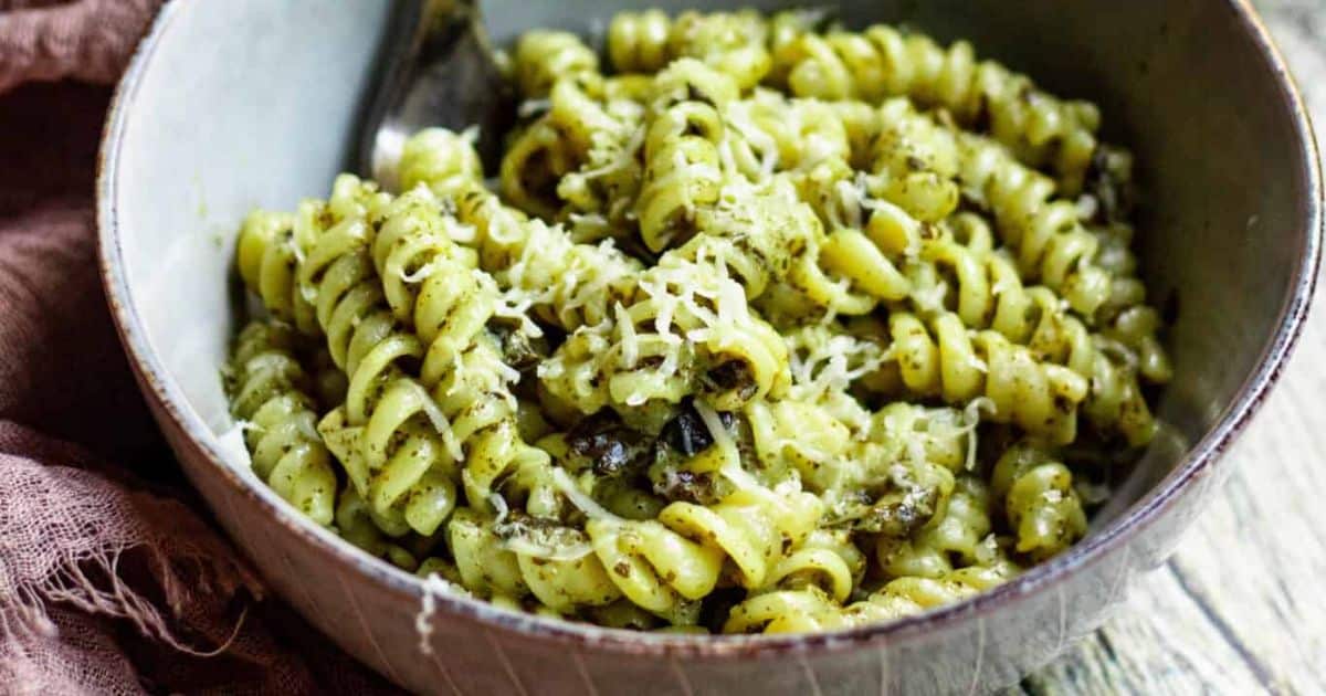 A bowl of rotini pasta with pesto sauce and grated cheese on top, with a spoon inside the bowl.