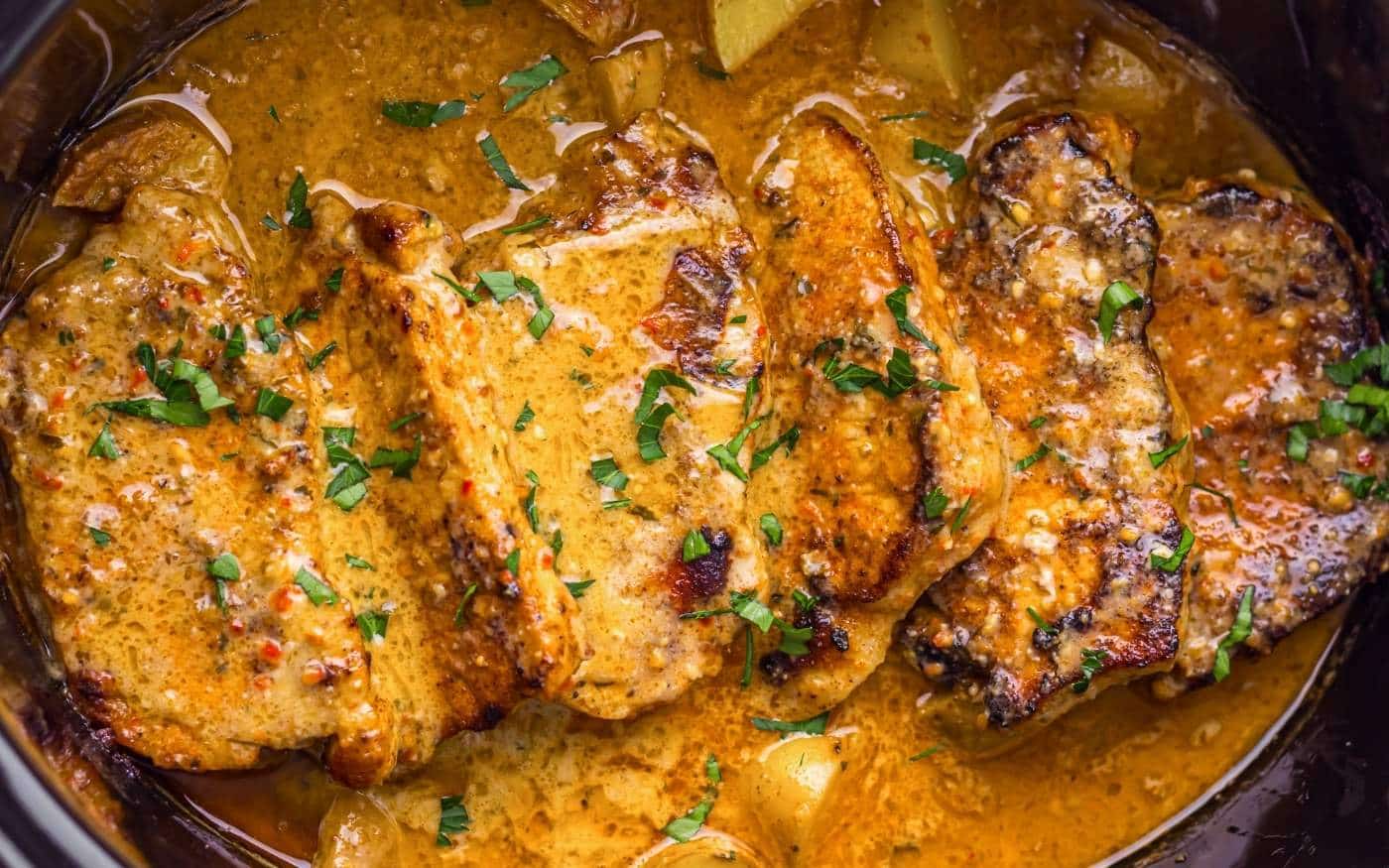 Four pork chops in creamy sauce, garnished with chopped parsley in a slow cooker.
