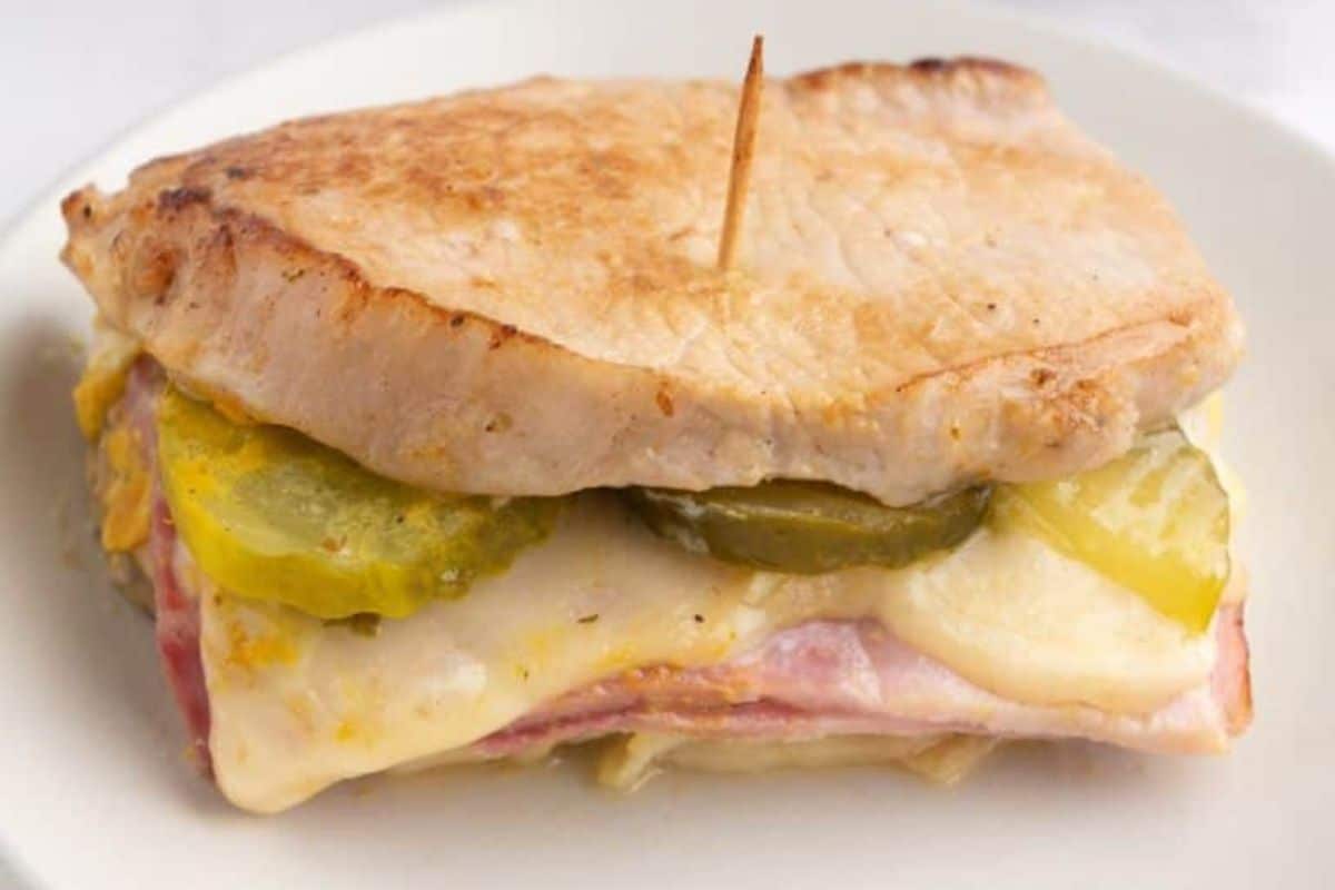 A sandwich with pork, melted cheese, ham, and pickles, held together with a toothpick on a white plate.