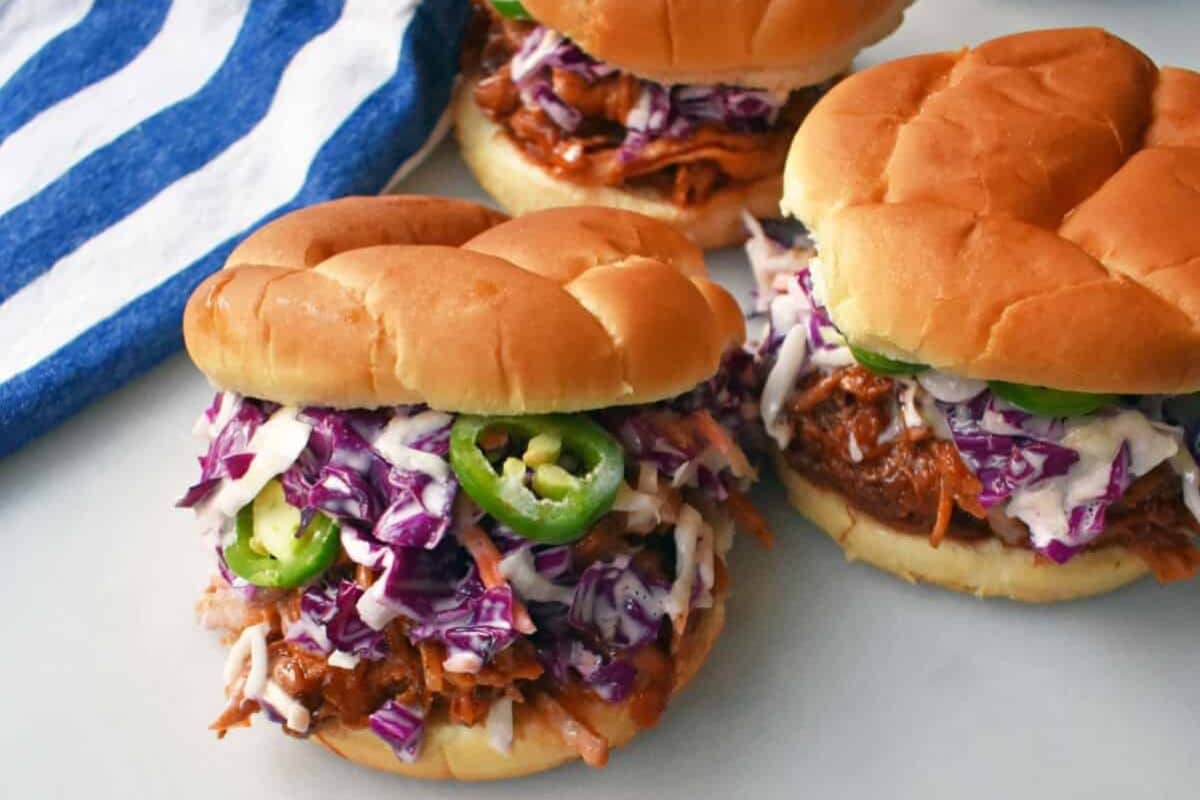 Three pulled pork sandwiches topped with purple coleslaw and jalapeño slices on buns, next to a blue striped towel.