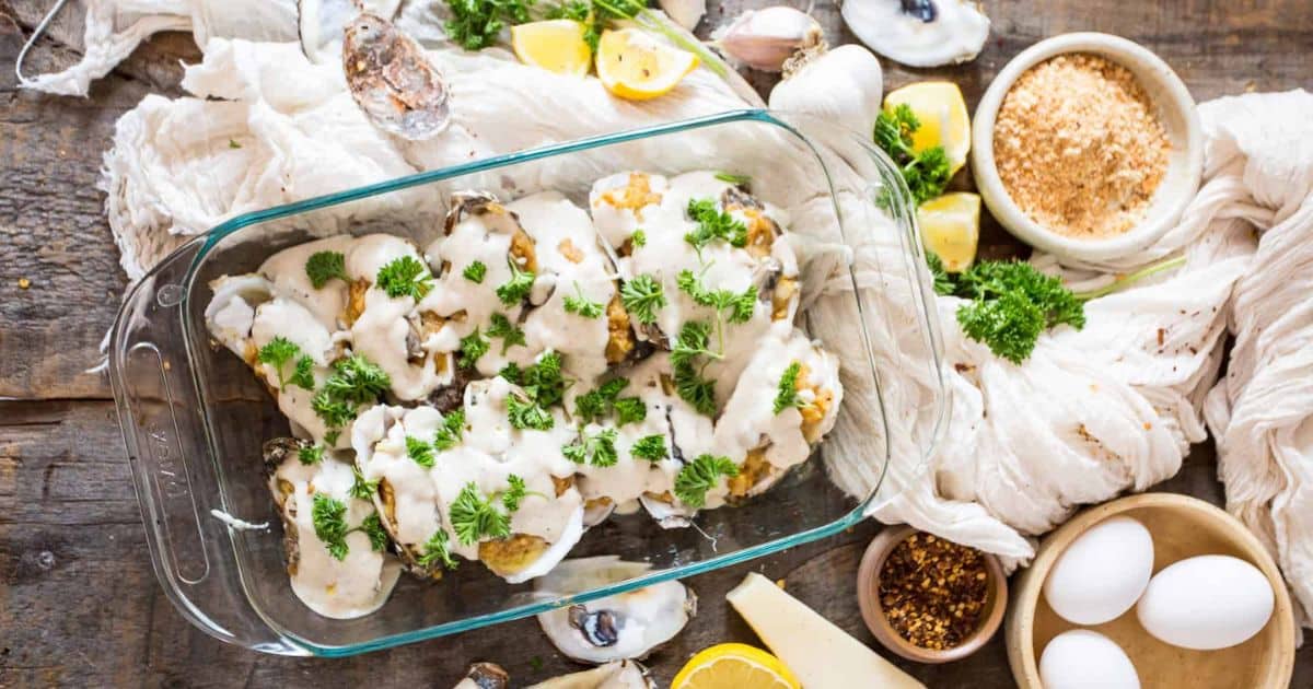 Glass dish of baked oysters topped with creamy sauce and parsley, surrounded by ingredients on a rustic table.