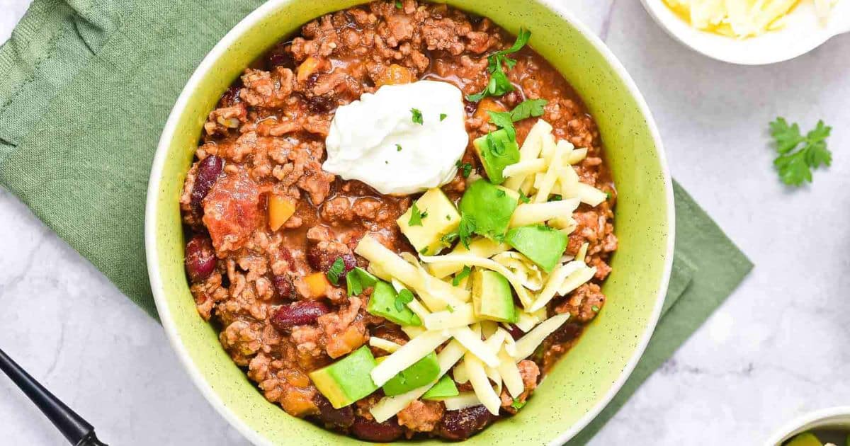 A bowl of chili topped with shredded cheese, avocado, and sour cream, on a green napkin.