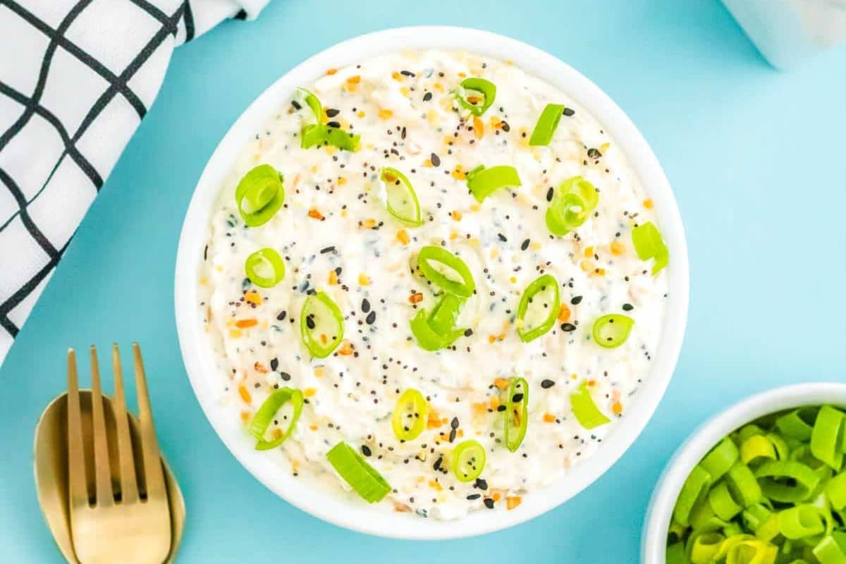 Bowl of creamy dip topped with chopped green onions and seeds on a blue surface with gold utensils nearby.