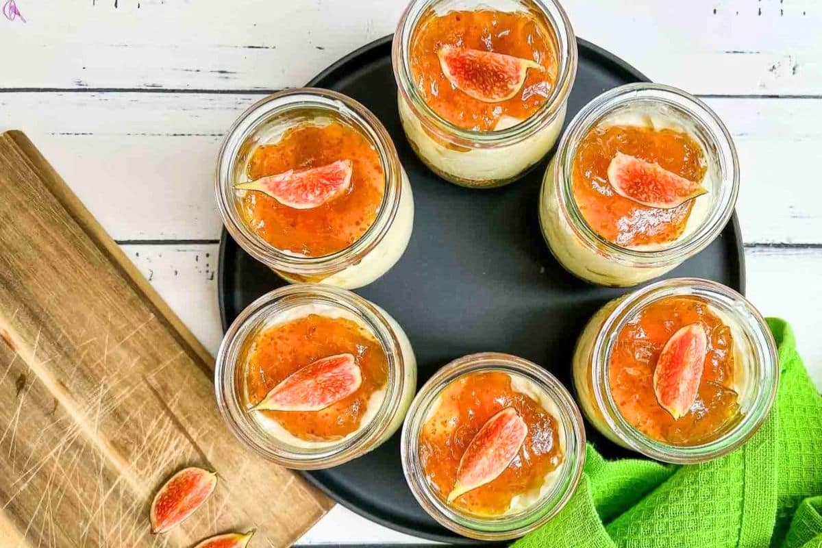 Seven dessert jars topped with fig jam and fresh fig slices on a black plate, next to a green cloth.