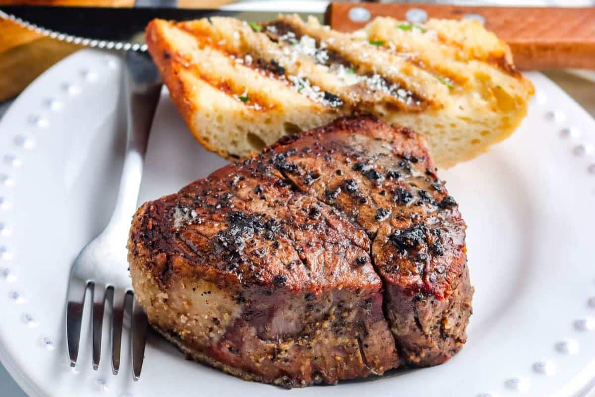 A grilled filet mignon steak and two slices of toasted bread on a white plate with a fork and knife.