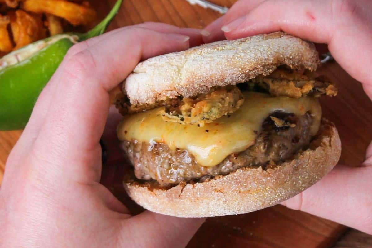 Hands holding a cheeseburger on an English muffin with fried pickles and melted cheese.