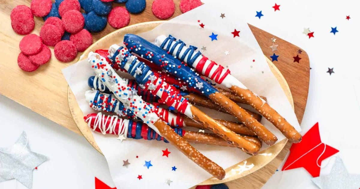 Pretzel rods dipped in red, white, and blue chocolate with sprinkles on a plate, surrounded by star confetti.