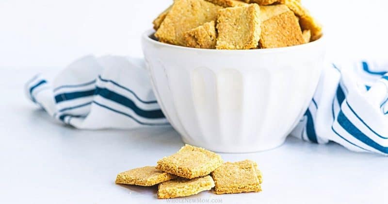 Square homemade crackers in a white bowl with a striped cloth and a few crackers placed in front.