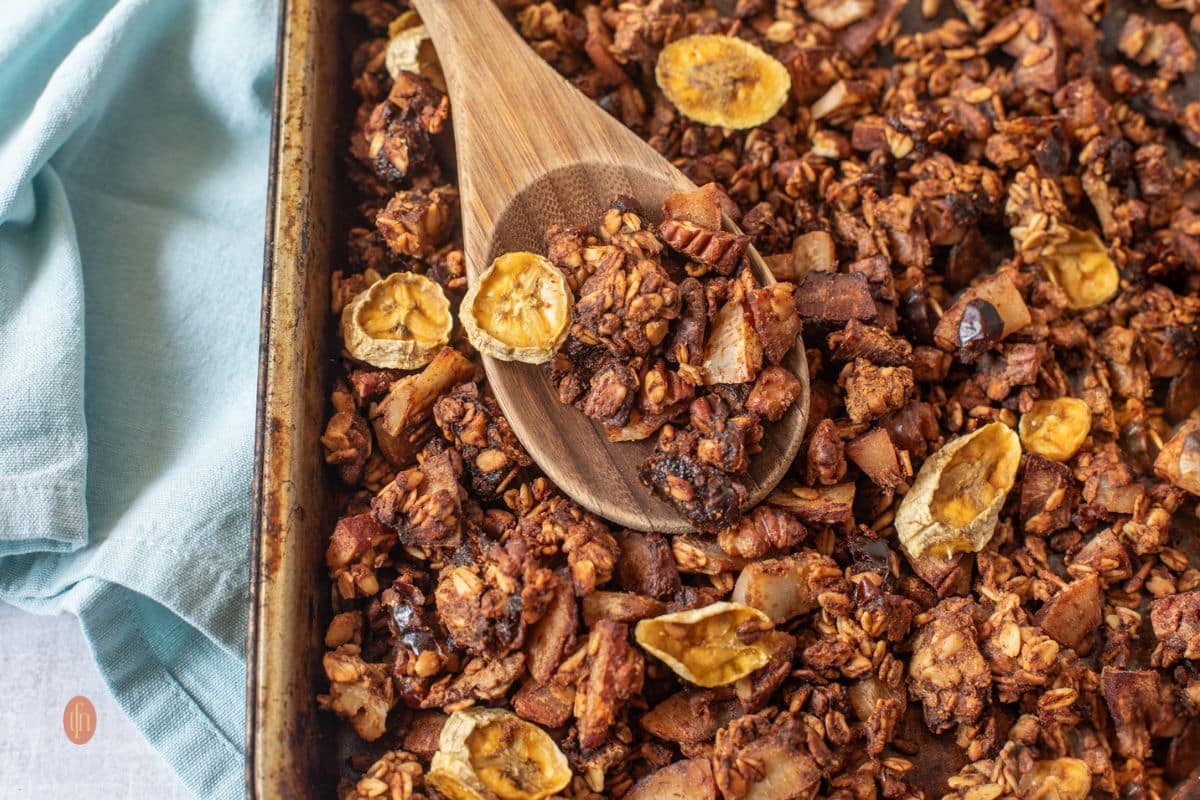 A baking tray of granola with banana chips, nuts, and a wooden spoon, next to a light blue cloth.
