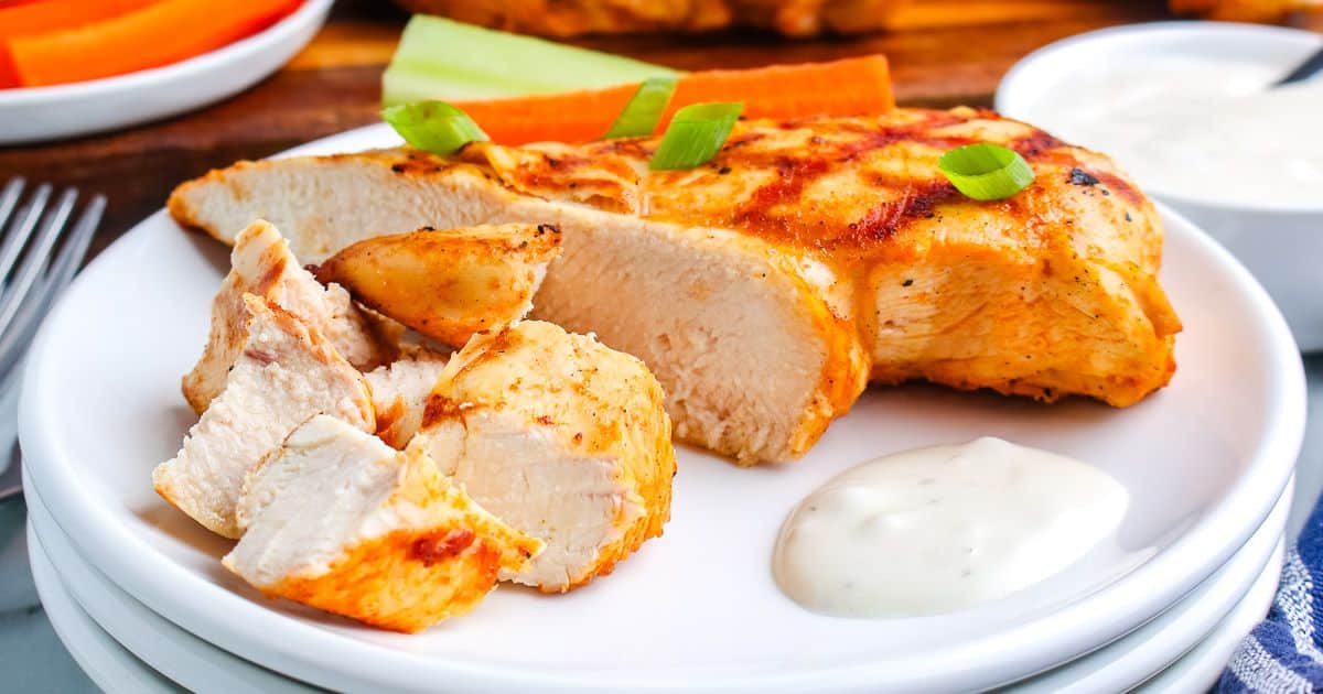Grilled chicken breast sliced on a white plate with ranch sauce, carrot, celery, and green onion garnish.