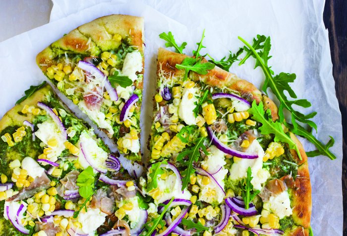 Vegetable pizza topped with arugula, corn, red onion, cheese, and pesto on a parchment-lined surface.