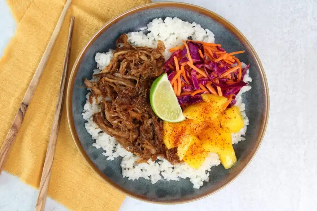 A bowl of rice topped with shredded meat, slaw, mango slices, and a lime wedge; chopsticks beside the bowl.