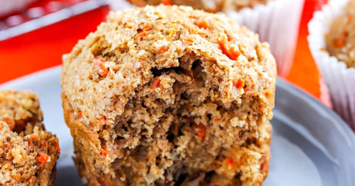 A close-up of a carrot muffin with a bite taken out, showing its moist, textured interior on a plate.