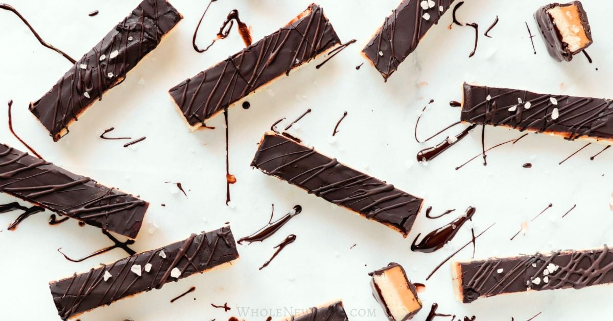 Rectangular chocolate-covered bars with caramel filling on a white surface, drizzled with chocolate and sea salt.