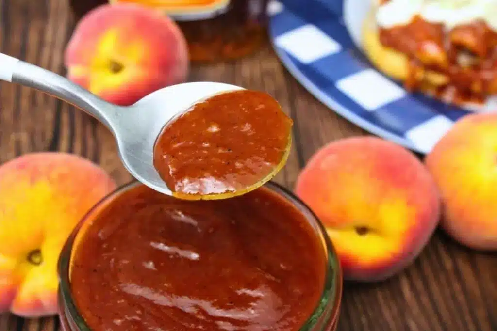 A spoonful of peach BBQ sauce held over a jar, with fresh peaches and food in the background.