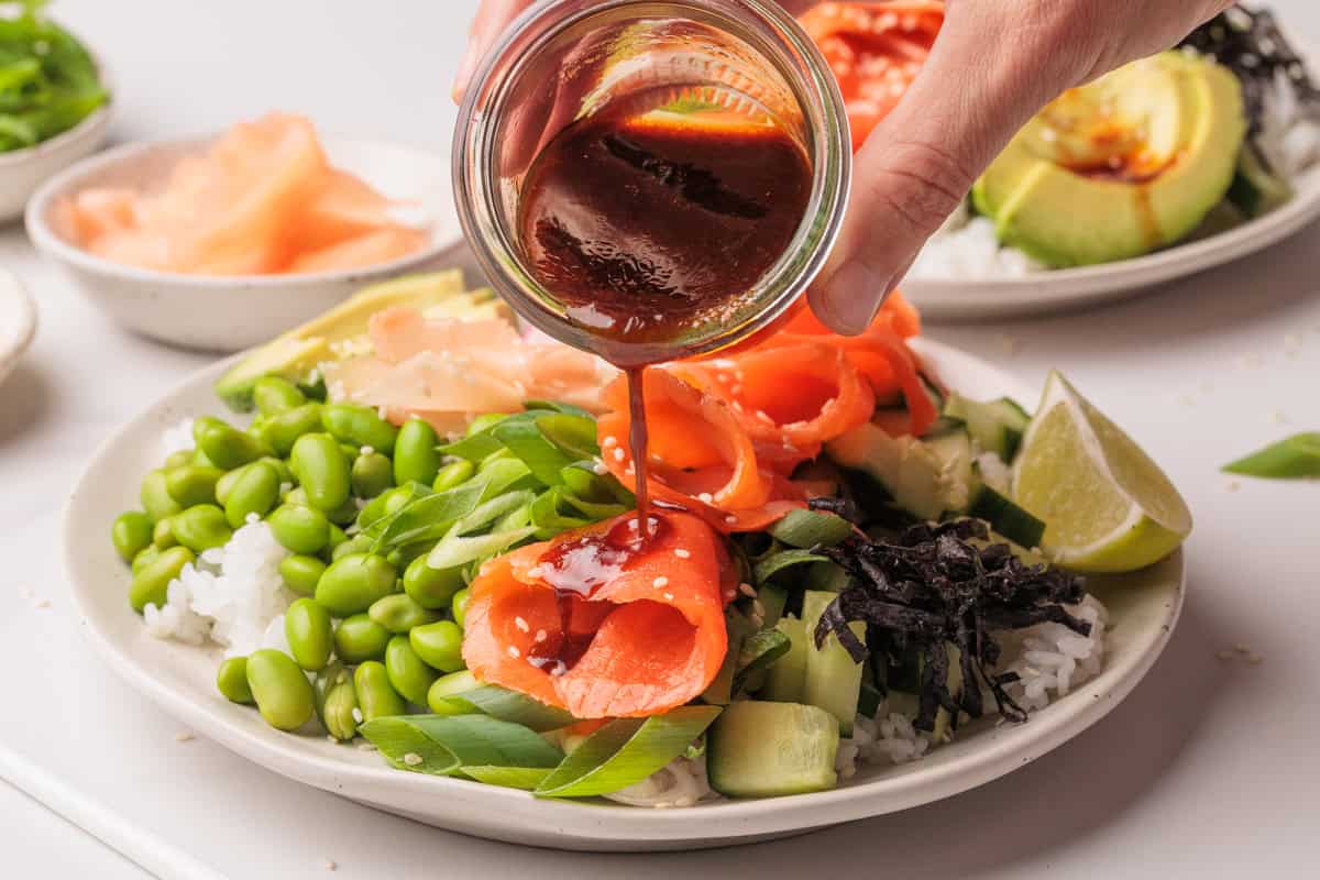 A hand pours sauce over a poke bowl with salmon, edamame, rice, cucumber, seaweed, and lime.