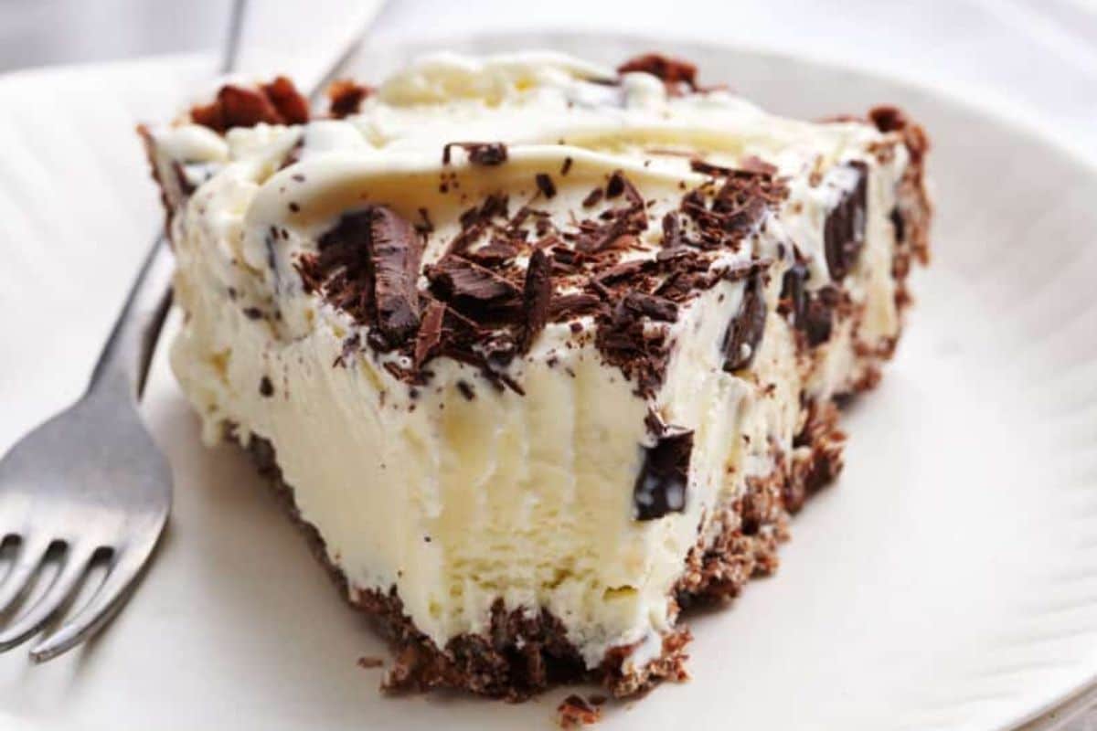 A slice of creamy ice cream pie with chocolate shavings and a chocolate crust sits on a white plate with a fork.