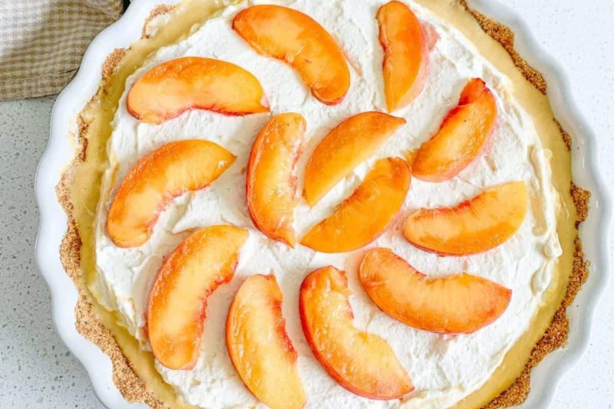 A cream pie topped with whipped cream and fresh peach slices in a white pie dish.