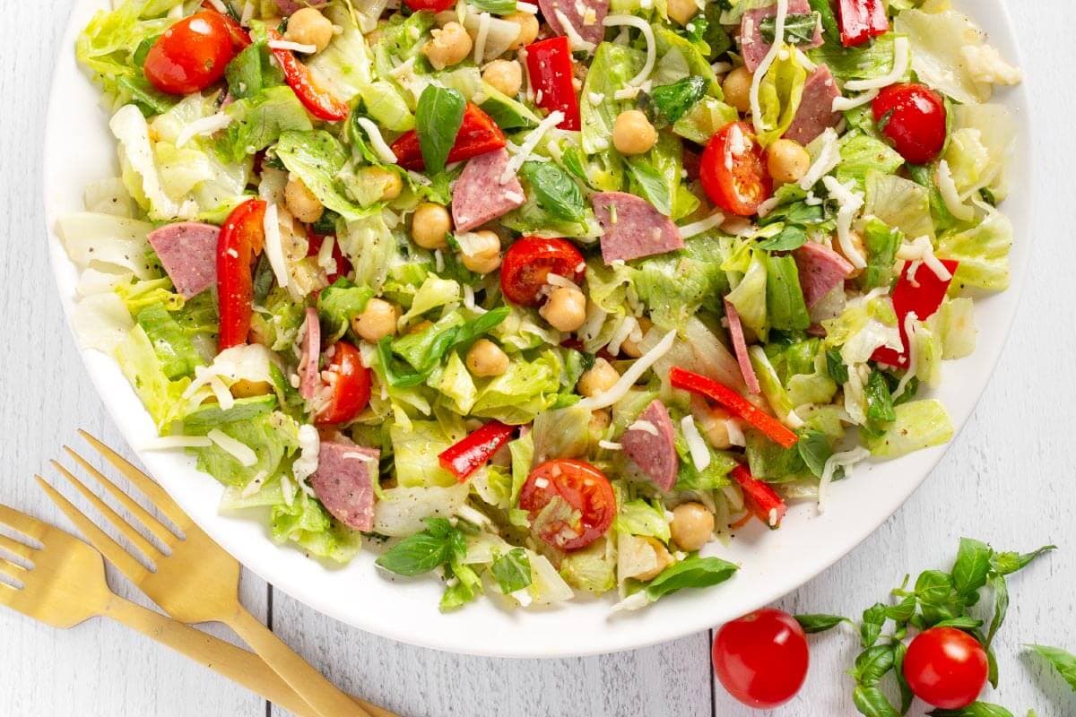 A bowl of colorful salad with lettuce, tomatoes, chickpeas, salami, red peppers, and grated cheese.