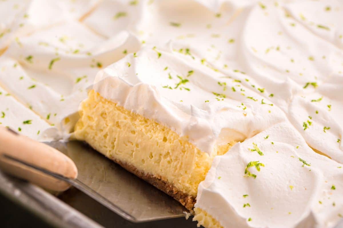 A creamy, whipped-topped dessert bar being lifted from a pan, garnished with fresh lime zest.