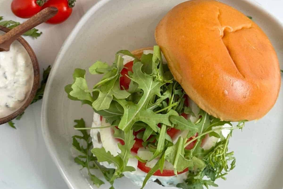 A sandwich with arugula, tomato, and sauce on a bun, served on a white plate with sauce and tomatoes nearby.