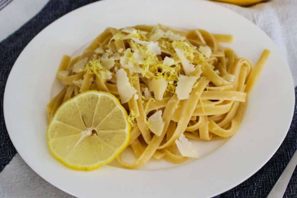 Fettuccine pasta with grated cheese and lemon zest, garnished with a lemon slice on a white plate.