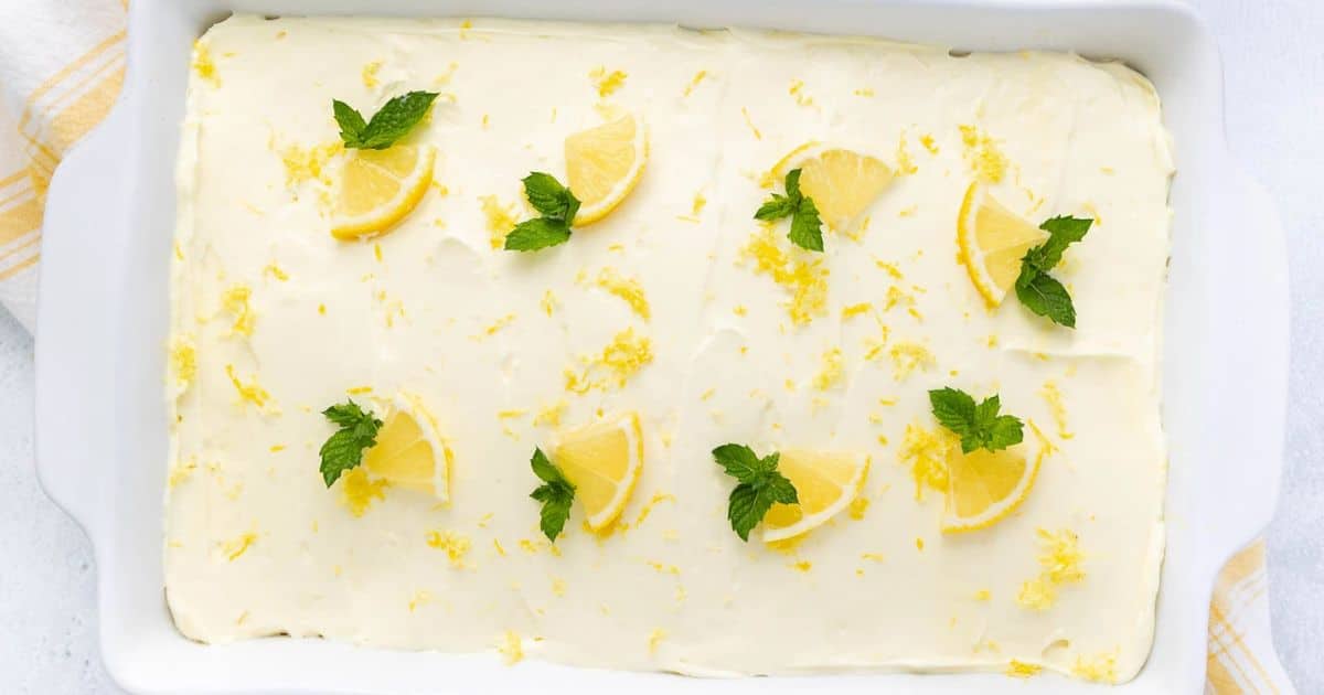 Rectangular lemon sheet cake with white frosting, lemon slices, zest, and mint leaves on top in a white dish.