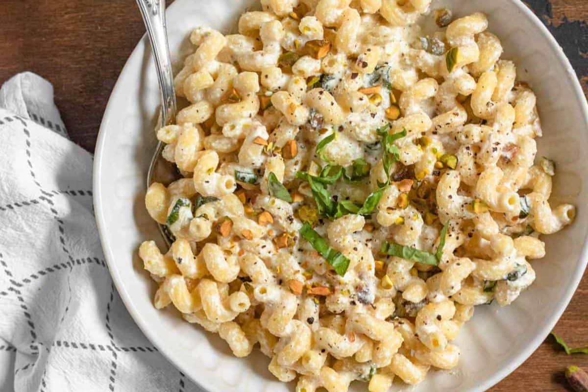 Bowl of creamy pasta topped with chopped herbs and nuts, with a fork and a checkered napkin beside it.