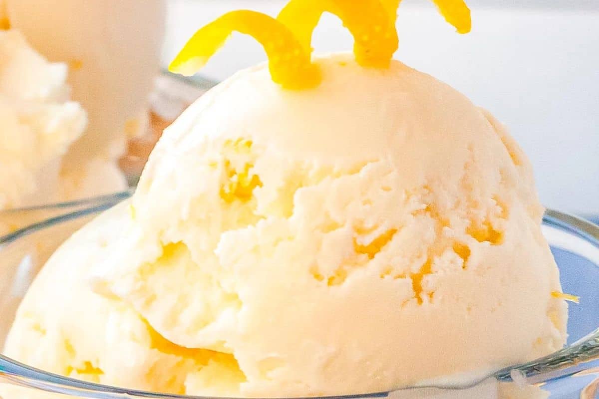 A scoop of creamy lemon ice cream in a glass bowl, garnished with lemon peel twists on top.