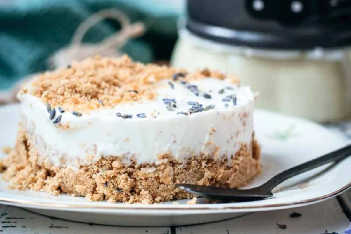 A slice of creamy cheesecake with a crumbly crust, topped with lavender and served on a plate with a fork.