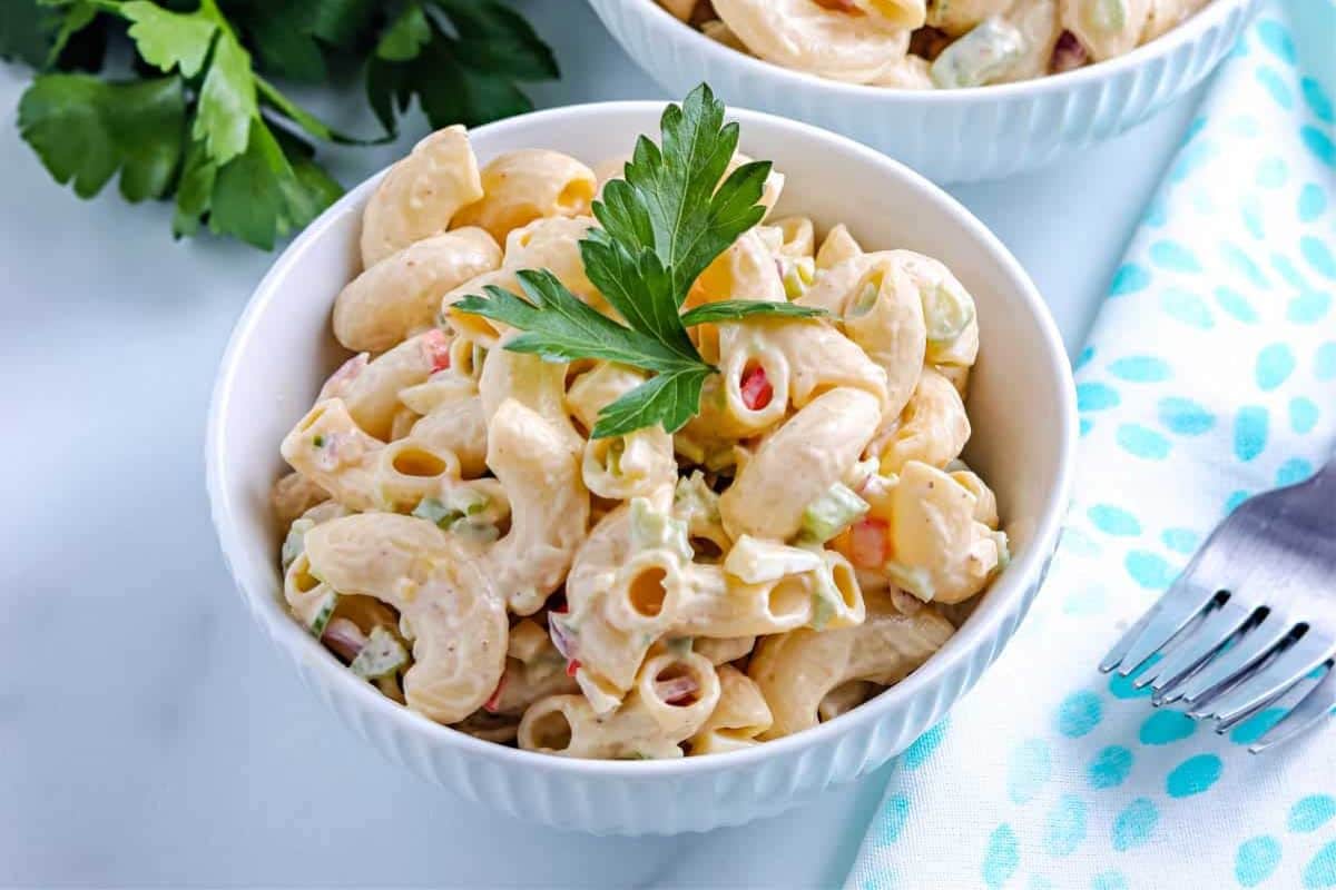 A white bowl of creamy macaroni salad garnished with parsley, next to a fork and a blue napkin.