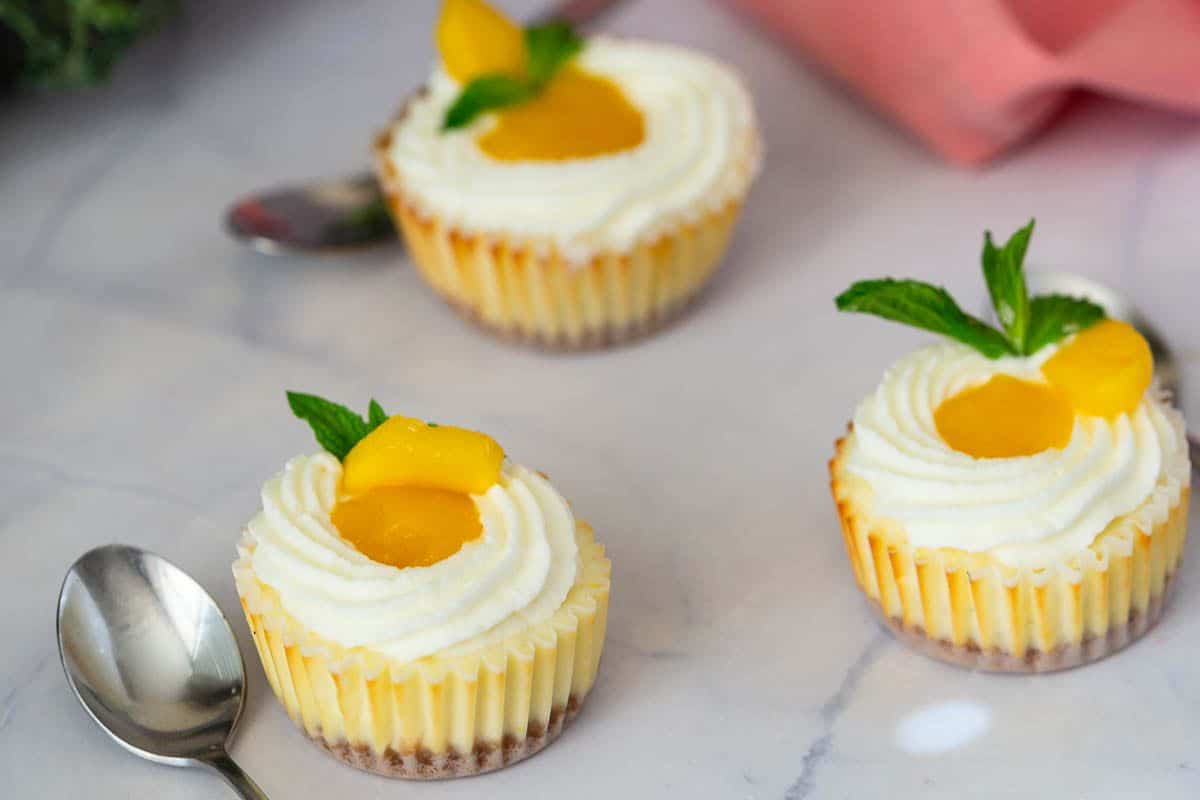 Three mini cheesecakes with whipped cream, peach slices, and mint leaves on a marble surface with two spoons.