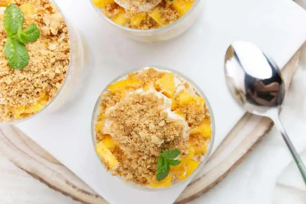Top view of dessert cups with crumbled topping, mango chunks, whipped cream, and mint leaves beside a spoon.