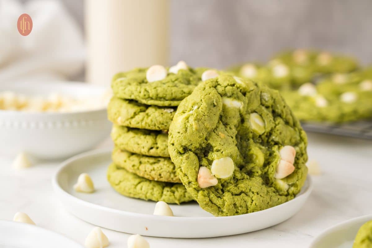 A stack of green cookies with white chocolate chips on a white plate, with more cookies in the background.